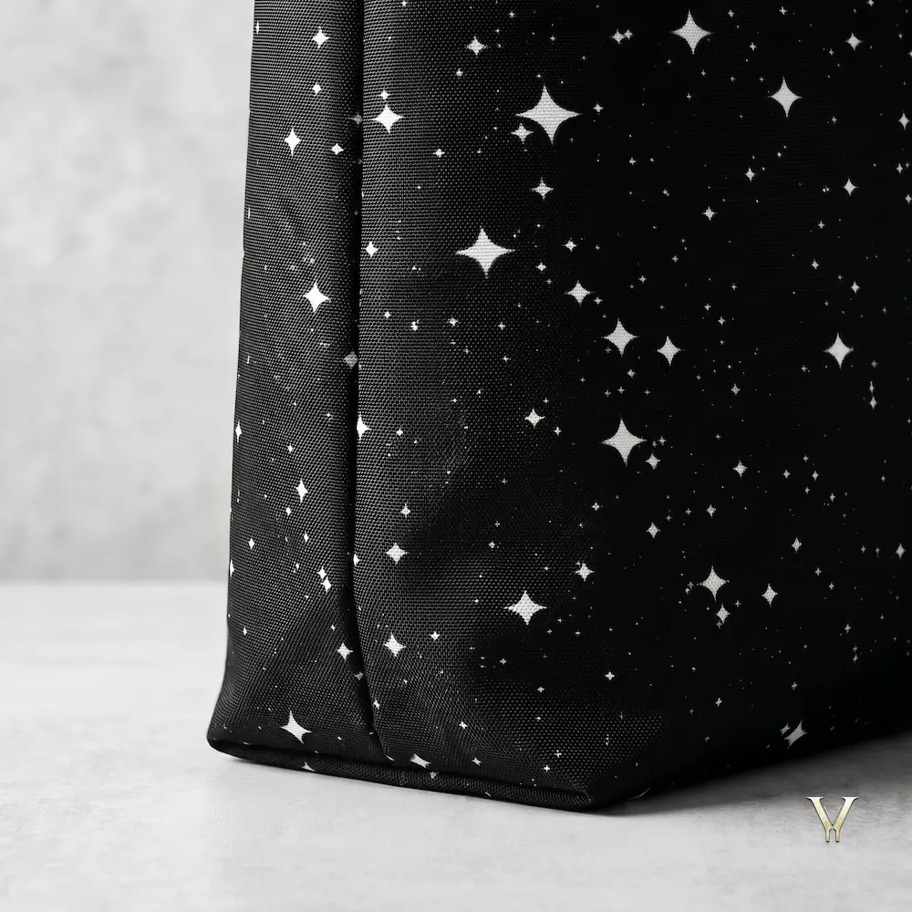 As shown in Close-Shot Square.webp, a detailed macro, square-cropped close-up highlights the lower corner and sturdy textured fabric of a black Designer Larger Tote Bag. The material features a seamless, high-contrast print of white four-pointed stars and cluster dots resembling a night sky constellation, resting on a smooth grey surface.