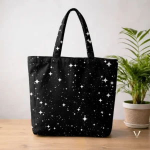 A large black Designer Tote Bag with an all-over print of white four-pointed stars and tiny dots mimicking a night sky, displayed on a wooden surface next to a potted green plant.