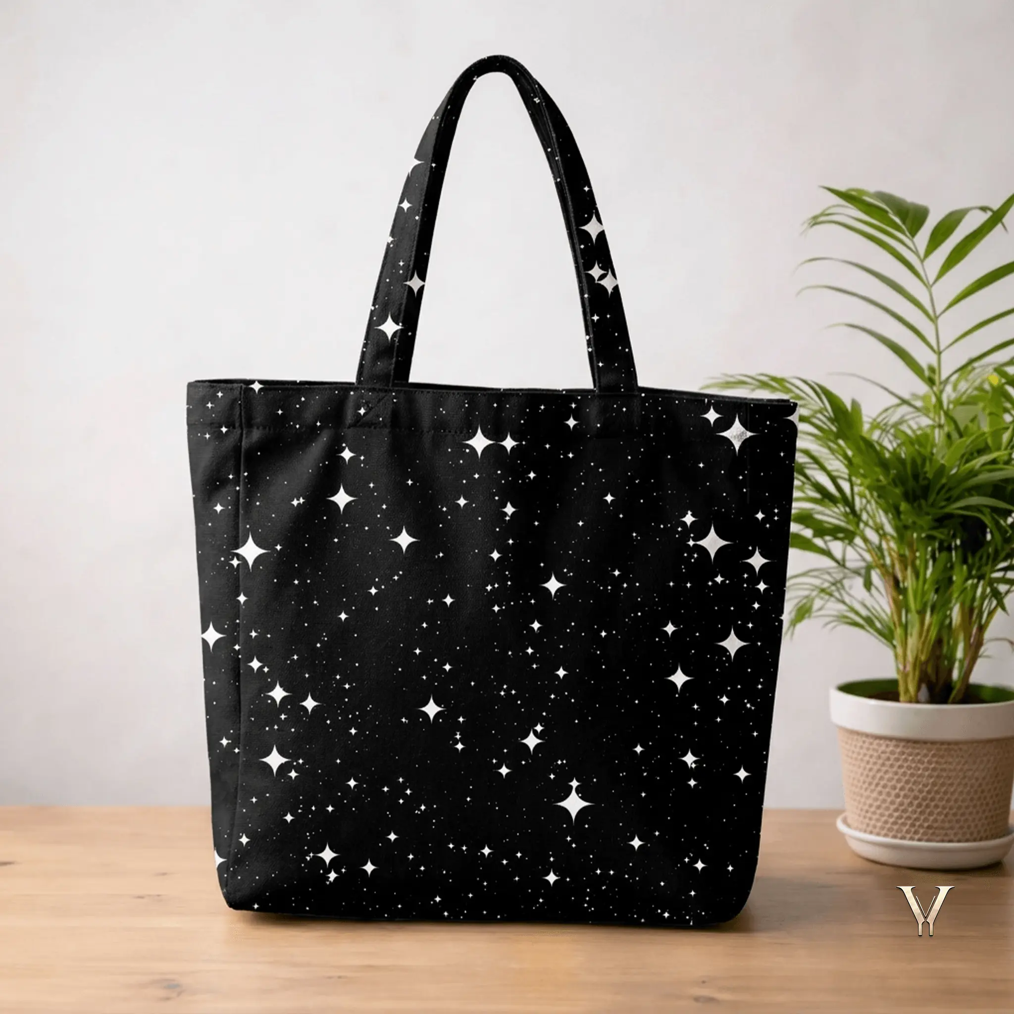 A large black Designer Tote Bag with an all-over print of white four-pointed stars and tiny dots mimicking a night sky, displayed on a wooden surface next to a potted green plant.