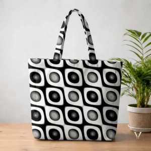 As displayed in Eco Sphere Tote Bag Mock up.webp, a large Designer tote bag featuring an all-over abstract geometric pattern in black, white, and grey is positioned on a wooden tabletop. The pattern consists of repeating, seamless square blocks containing rounded, 3D-effect spherical shapes that create an optical illusion. A terracotta potted green plant sits in the background against a light grey wall.